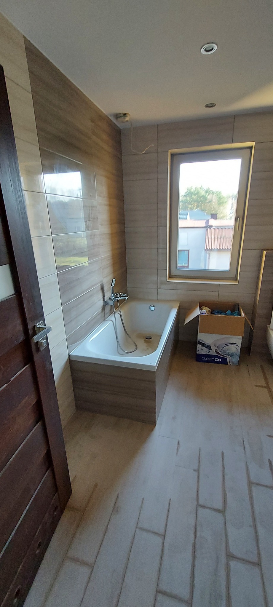 Nowa łazienka z wanną obudowaną płytkami imitującymi drewno, jasnymi płytkami na ścianach, oknem z widokiem na domy i podłogą w trakcie układania z widocznymi fugami.