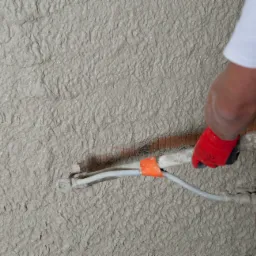 Nakładanie szarego tynku maszynowego na ścianę z widoczną instalacją elektryczną, robocze rękawice ochronne na dłoniach wykonawcy.