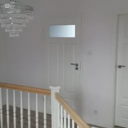 Jasne wnętrze z trzema białymi drzwiami wewnętrznymi, kryształowym żyrandolem i drewnianymi balustradami. Jedne drzwi z przeszkleniem.