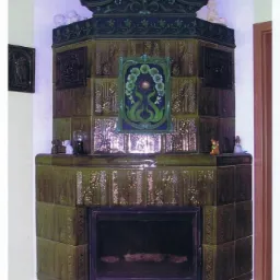 Rustykalny, zielony kominek kaflowy z ozdobnym zwieńczeniem i motywami roślinnymi. Na półce dekoracje. Widoczne detale płytek i konstrukcji kominka.