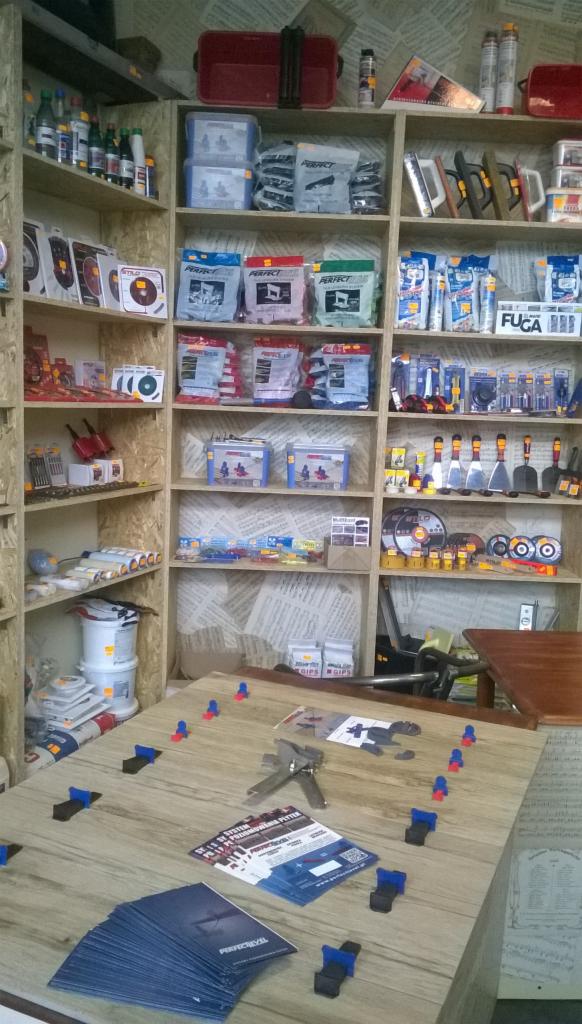 Sklep z artykułami budowlanymi: półki wypełnione narzędziami, chemią budowlaną i akcesoriami do układania płytek. Na stole ulotki i system poziomowania płytek.