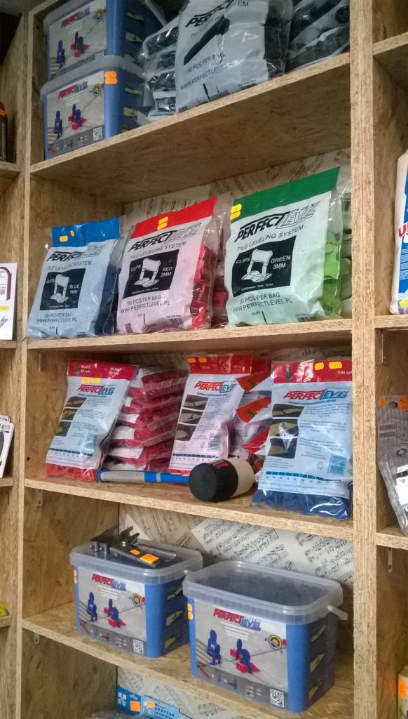 Regał z systemami poziomowania płytek Perfect Level w kolorowych opakowaniach i pojemnikach, narzędzia glazurnicze, ekspozycja w sklepie.