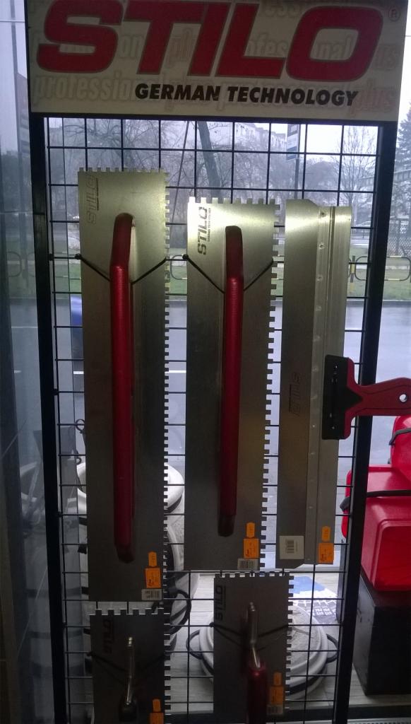 Ekspozycja w sklepie: trzy długie, metalowe pace z czerwonymi uchwytami marki Stilo, wiszące na czarnej kratownicy na tle okna i logo firmy.