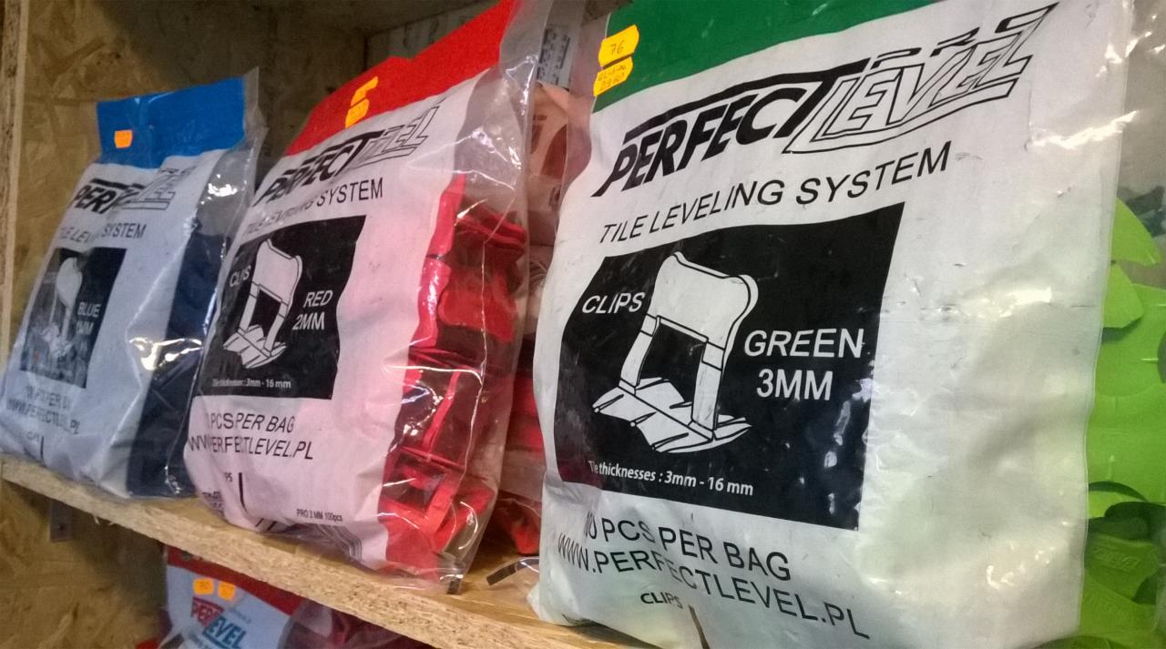 Półka z paczkami systemu poziomowania płytek Perfect Level w kolorach: niebieskim, czerwonym i zielonym. Widoczne klipsy 2mm i 3mm. Produkty do układania płytek.