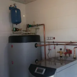 Szary zasobnik ciepłej wody użytkowej z niebieskim naczyniem wzbiorczym i miedzianymi rurami w pomieszczeniu z białymi płytkami i mozaiką.