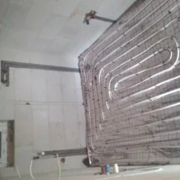 Montaż ogrzewania podłogowego: białe rurki ułożone w pętle na siatce w surowym wnętrzu, widoczne elementy mocujące i instalacja na suficie.