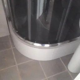 Fragment wykończonej łazienki z toaletą, umywalką na postumencie i kabiną prysznicową z chromowanymi detalami, widoczna szczotka do sprzątania na szarej podłodze z płytek.