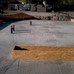 Betonowa płyta fundamentowa z wystającymi rurami i drewnianym szalunkiem, przygotowana pod budowę. Widoczne zbrojenie w tle.