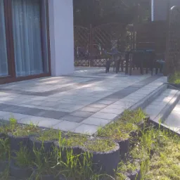 Ułożona kostka brukowa w dwóch odcieniach szarości tworząca wzór na tarasie przed domem, z obrzeżem z palisady betonowej i schodami z kostki, w tle meble ogrodowe i drewniana altana.