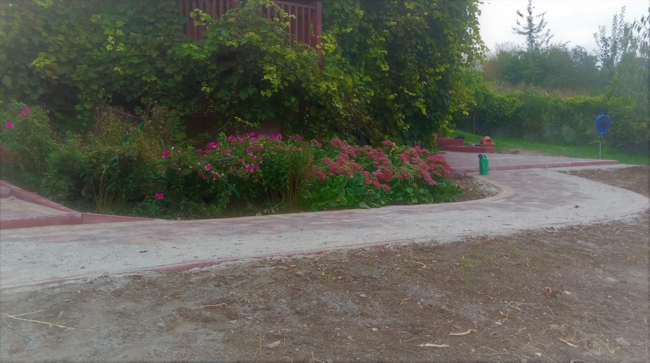 Zakrzywiony chodnik z szarej kostki z czerwoną obwódką otoczony zielenią, kwiatami i drzewami w tle.