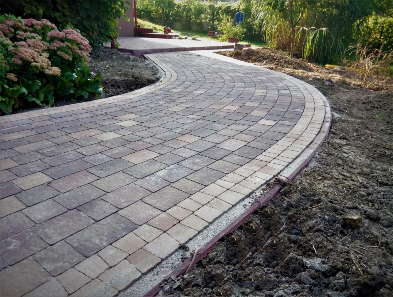 Zakrzywiony chodnik z kostki brukowej w odcieniach szarości i brązu, obrzeża z czerwonej kostki, widoczne ślady świeżo ułożonej ziemi po bokach, w tle roślinność ogrodowa.