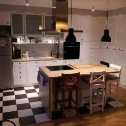 Jasna kuchnia z wyspą, czarno-białą podłogą w szachownicę i drewnianym blatem, oświetlona czarnymi lampami wiszącymi.