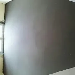 Gładka, szara ściana w rogu pokoju, widoczna obok okna z surowym wykończeniem. Minimalistyczne wnętrze w trakcie prac wykończeniowych.