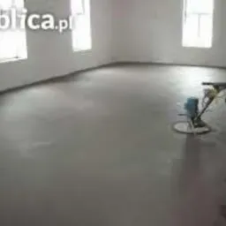 Szara, gładka posadzka w jasnym pomieszczeniu. Widoczna maszyna do polerowania podłóg i ślady świeżo wykonanej wylewki przy ścianie. Minimalistyczne wnętrze.