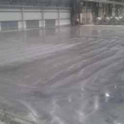 Świeżo wylana, gładka powierzchnia podłogi w hali przemysłowej, widoczne ślady po rozprowadzaniu materiału, w tle rolki blachy i konstrukcja hali.