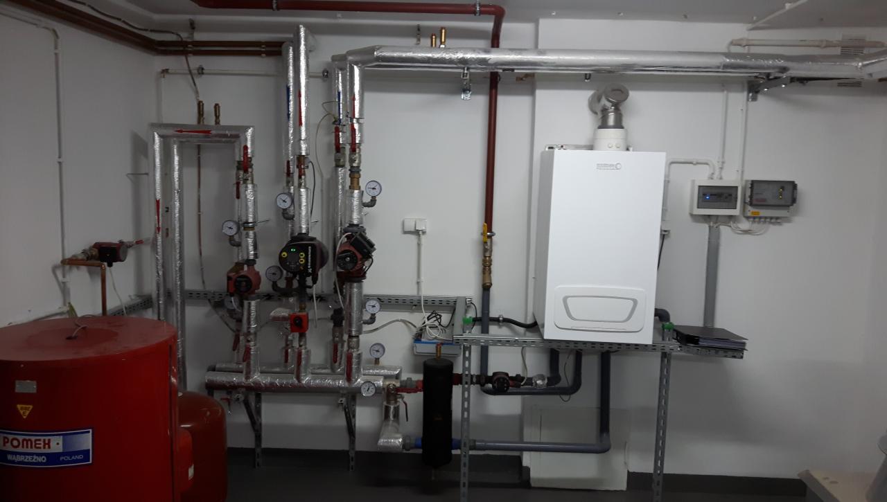 Nowoczesny, biały piec gazowy zamontowany na metalowym stelażu, obok rozbudowana instalacja z rur, zaworów, manometrów i pomp obiegowych, całość w pomieszczeniu technicznym z białymi ścianami.