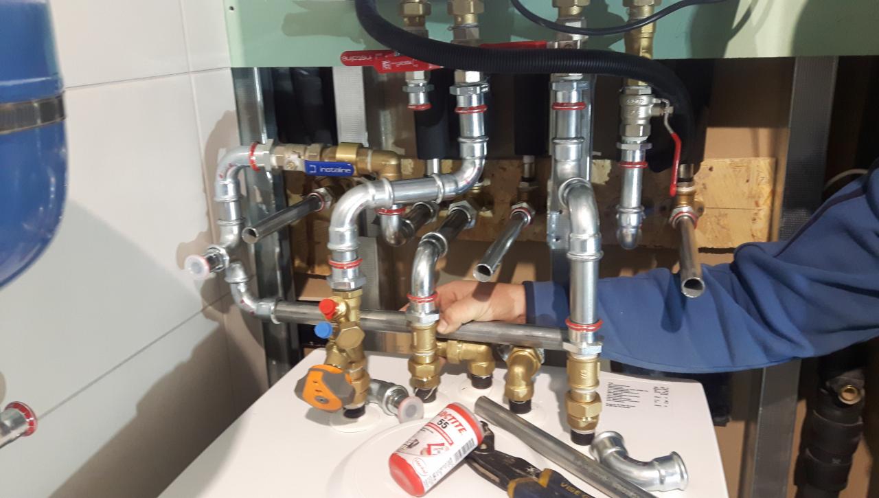 Instalacja grzewcza w trakcie montażu: widoczne rury miedziane, zawory z czerwonymi uchwytami, kształtki, puszka Loctite 55 oraz narzędzia monterskie. Dłoń w niebieskiej bluzie przytrzymuje rurę.