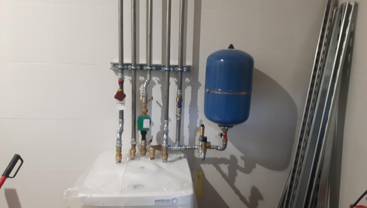 Nowy piec centralnego ogrzewania w folii ochronnej, z podłączonymi rurami, zaworami i niebieskim zbiornikiem wyrównawczym, widoczne także profile montażowe.