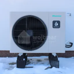 Biała zewnętrzna jednostka pompy ciepła Hisense Hi-Therma, zamontowana na betonowych blokach w zimowym otoczeniu z resztkami śniegu.