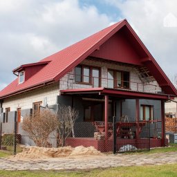 WOJTMAR - Dajemy Energię! - Dom jednorodzinny z czerwoną dachówką, częściowo ocieplony szarym styropianem, z widocznymi panelami słonecznymi na dachu sąsiedniego budynku. Na pierwszym planie piasek i ogrodzenie z siatki.