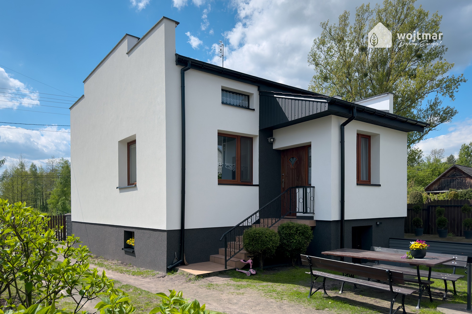 Nowoczesny, dwukondygnacyjny dom po termomodernizacji z białymi ścianami, grafitowym cokołem i ciemną blachą na dachu, z logo firmy wykonującej prace w prawym górnym rogu, otoczony zielenią.