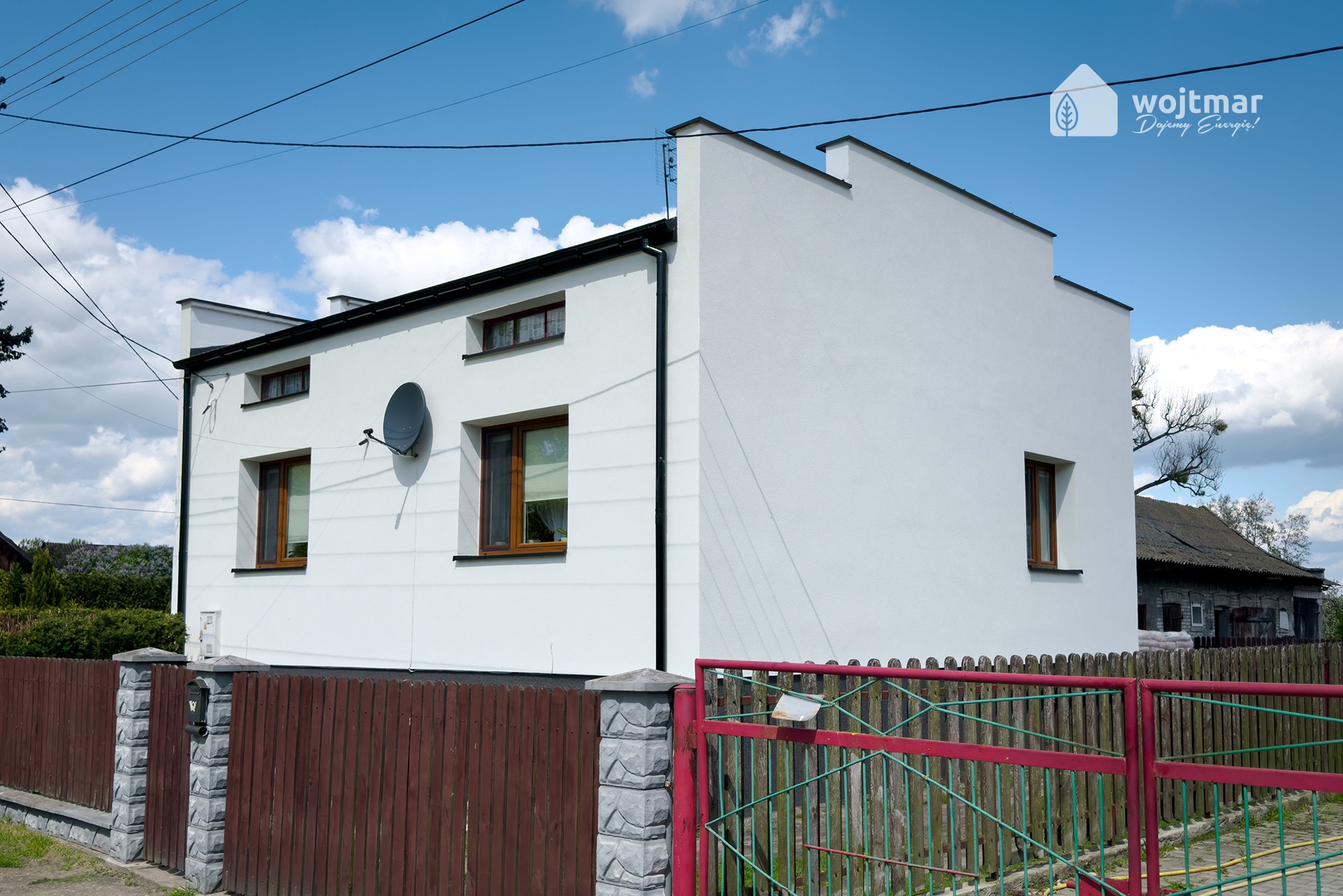 Biały, dwukondygnacyjny dom jednorodzinny z brązowymi oknami i anteną satelitarną, widziany zza brązowo-czerwonego ogrodzenia. W prawym górnym rogu widoczne logo firmy Wojtmar.