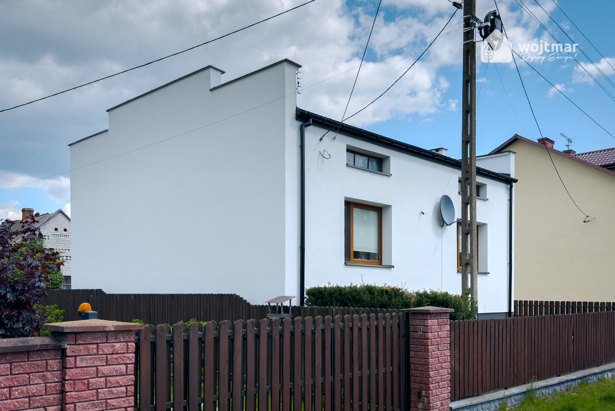 Biały, otynkowany dom jednorodzinny z brązową stolarką okienną i anteną satelitarną na elewacji, ogrodzony drewnianym płotem z ceglanymi słupkami, widoczny słup energetyczny z przewodami.
