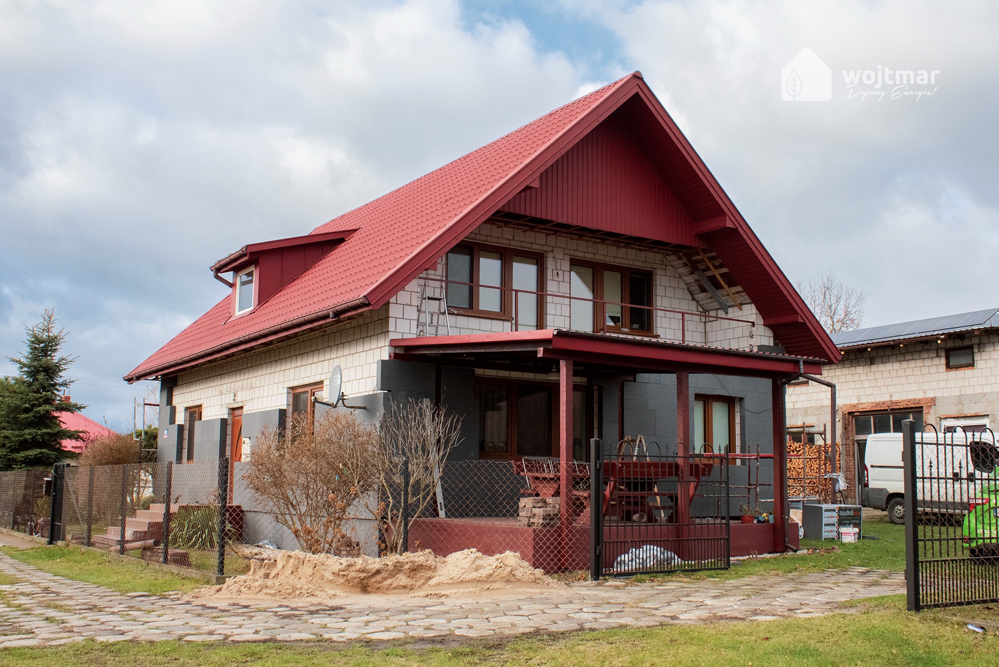 Dom jednorodzinny z czerwoną dachówką, częściowo ocieplony szarym styropianem, z widocznymi panelami słonecznymi na dachu sąsiedniego budynku. Na pierwszym planie piasek i ogrodzenie z siatki.
