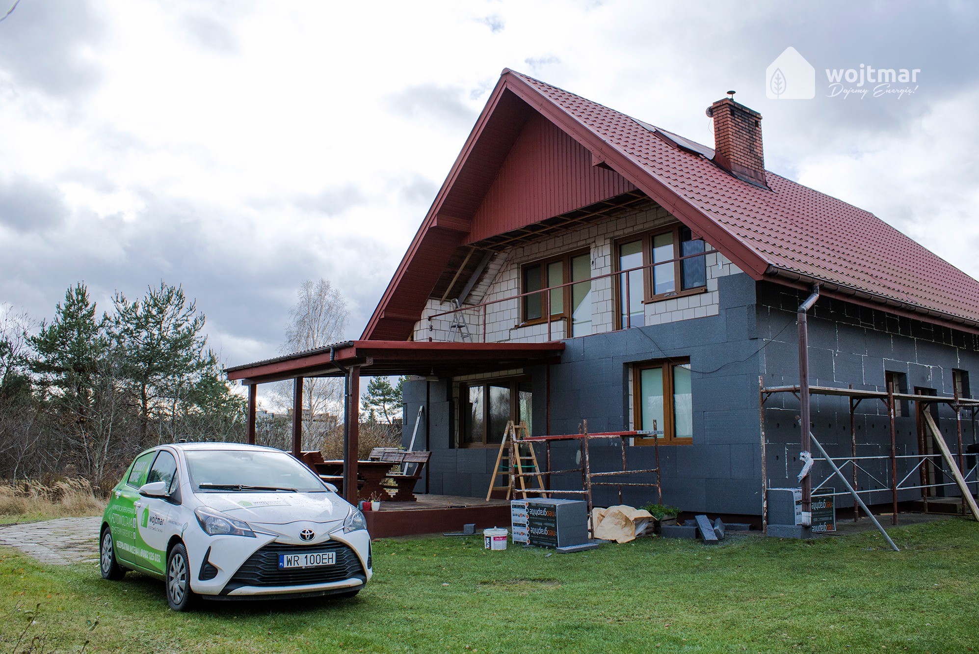 Dom jednorodzinny w trakcie ocieplania grafitowym styropianem, widoczne rusztowania, drabina i materiały budowlane, na pierwszym planie samochód firmowy z logo.