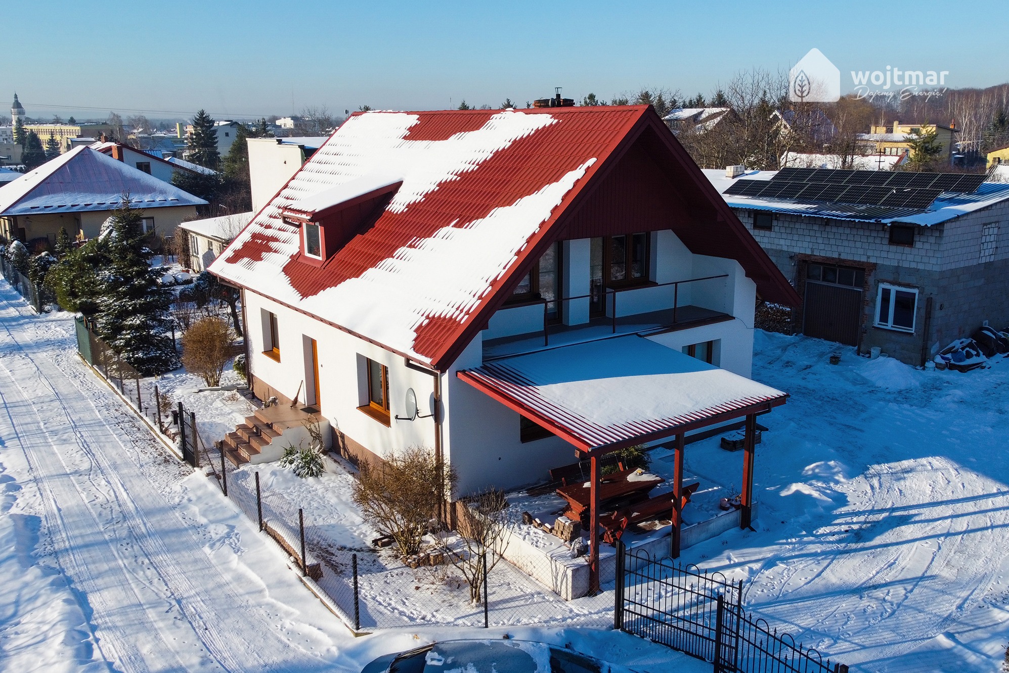 Dom jednorodzinny z czerwoną dachówką częściowo pokrytą śniegiem, panele słoneczne na budynku gospodarczym w tle, zimowy krajobraz.