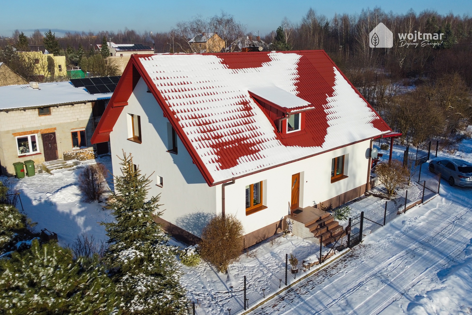 Dom jednorodzinny z czerwoną dachówką częściowo pokrytą śniegiem, widok z góry, obok drugi budynek z panelami fotowoltaicznymi na dachu, na pierwszym planie ośnieżone drzewa iglaste, w tle las.