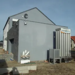 Nowoczesny dom z zainstalowaną zewnętrzną jednostką pompy ciepła, widoczna instalacja i teren w trakcie prac wykończeniowych.