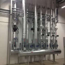 Rzędy izolowanych, metalowych rur z zaworami i manometrami w pomieszczeniu technicznym z białymi ścianami i betonową podłogą.