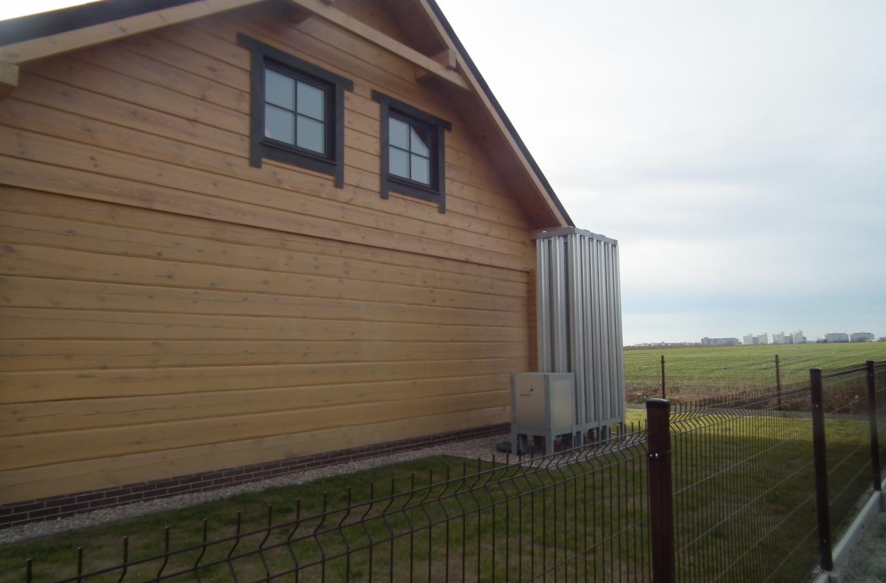 Zewnętrzna jednostka pompy ciepła powietrze-woda przy drewnianym domu, widok zza ogrodzenia, w tle pole i budynki.