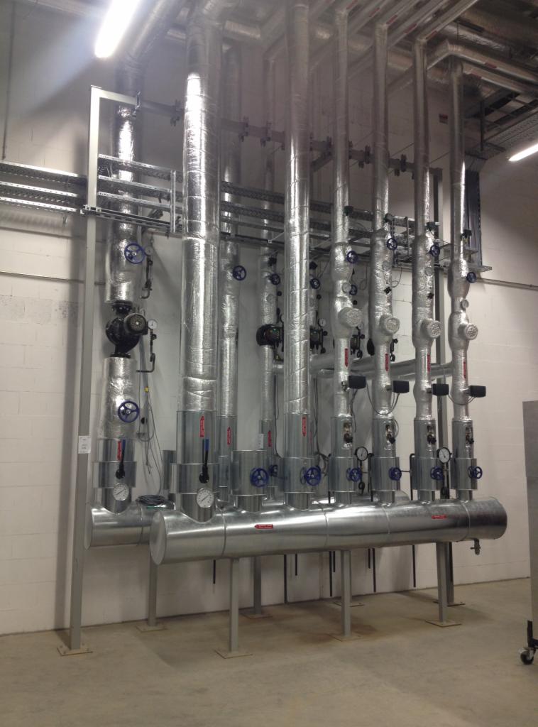 Rzędy izolowanych, metalowych rur z zaworami i manometrami w pomieszczeniu technicznym z białymi ścianami i betonową podłogą.