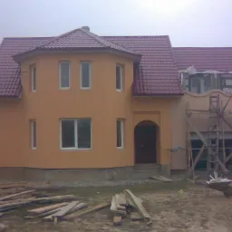 Dwukondygnacyjny dom z pomarańczową elewacją i ciemnoczerwoną dachówką, częściowo w rusztowaniach. Widoczne deski i taczka, sugerujące prace wykończeniowe.