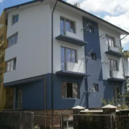 Dwukondygnacyjny dom z elewacją w odcieniach bieli i granatu, częściowo osłonięty siatką rusztowania. Widoczne balkony z betonowymi daszkami i elementy oświetlenia elewacyjnego.