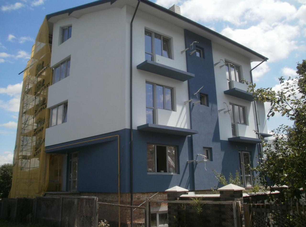 Dwukondygnacyjny dom z elewacją w odcieniach bieli i granatu, częściowo osłonięty siatką rusztowania. Widoczne balkony z betonowymi daszkami i elementy oświetlenia elewacyjnego.