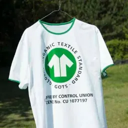 Biała koszulka z zielonym wykończeniem i nadrukiem certyfikatu GOTS, wisząca na wieszaku na tle zieleni.