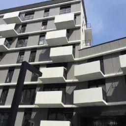 Nowoczesny budynek wielorodzinny z szarymi panelami elewacyjnymi i białymi, wystającymi balkonami o geometrycznych kształtach, widziany pod kątem z dołu, z czarną lampą uliczną w pierwszym planie.