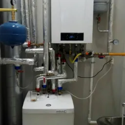 Nowoczesny piec gazowy dwufunkcyjny z podłączeniami w metalowej izolacji, widoczny zbiornik wyrównawczy i naczynie przeponowe, na górze pieca pusta szklanka.