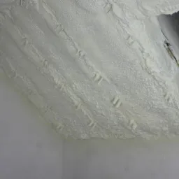 Biała, porowata warstwa izolacyjna na poddaszu w Miękini, z widocznymi elementami mocującymi i fragmentem otworu wentylacyjnego.