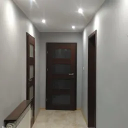 Korytarz z szarymi ścianami, trzema ciemnymi, drewnianymi drzwiami z przeszkleniami i jasną podłogą, oświetlony punktowymi lampami sufitowymi.