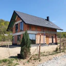 Surowy, dwukondygnacyjny dom z czerwonej cegły w trakcie budowy, z ciemną dachówką i czarnymi oknami, otoczony piaszczystym terenem i młodymi drzewkami.