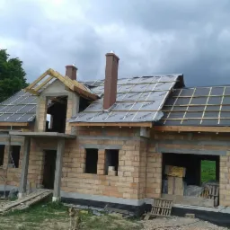 Budowa domu jednorodzinnego w trakcie krycia dachu membraną i łatami, widoczny surowy stan ścian z cegły, drewniana konstrukcja więźby dachowej i pies siedzący przed budynkiem.