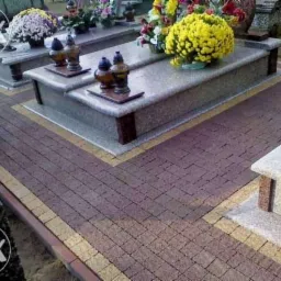 Granitowa kostka brukowa wokół nagrobków z kwiatami i lampionami, tworząca estetyczną, uporządkowaną przestrzeń na cmentarzu. Widok z poziomu gruntu.