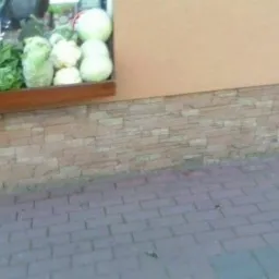 Elewacja budynku z dekoracyjną okładziną z imitacji kamienia i wystawą warzyw w oknie. Widoczna kostka brukowa na chodniku.