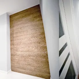 Wnętrze korytarza z panelami podłogowymi w odcieniach brązu, białą podłogą w części pomieszczenia, szarą ścianą i drewnianą okładziną na fragmencie ściany, białe drzwi z przeszkleniami.