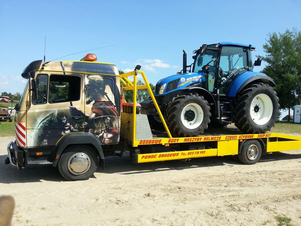 Laweta z grafiką przewozi niebieski traktor New Holland. Żółta platforma, widoczne numery telefonu pomocy drogowej. Tło: zielone drzewa, błękitne niebo.