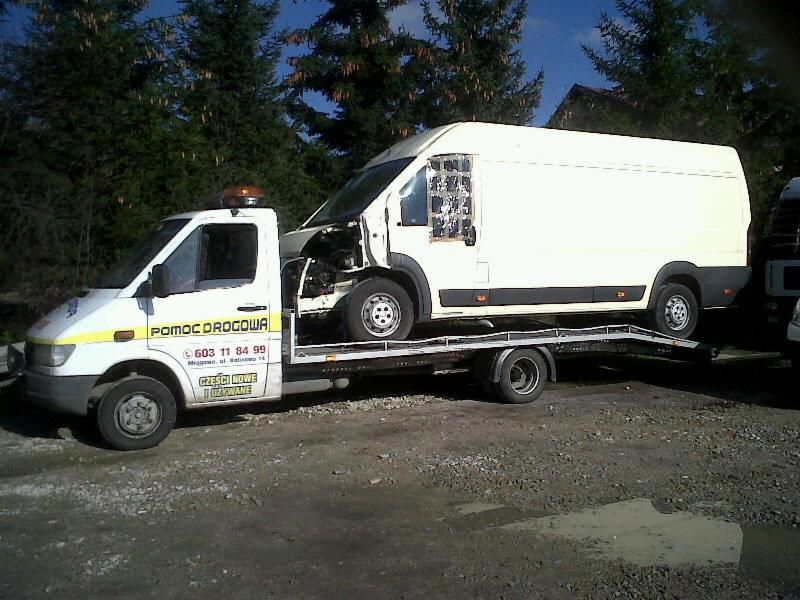 Laweta z białym busem na platformie, widoczne oznaczenia 'Pomoc Drogowa' i numer telefonu. Tło stanowią drzewa i budynek.